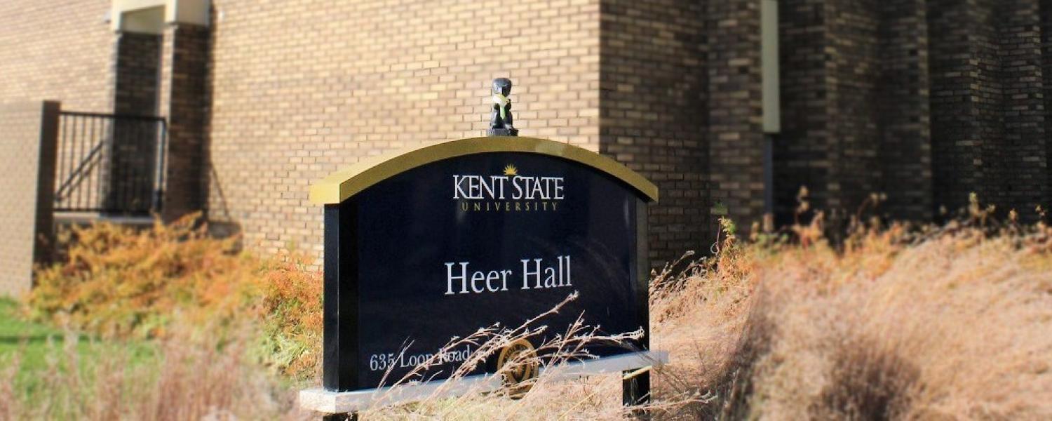 Black squirrel eating a piece of paper pie on top of the Heer Hall sign on Kent State's campus